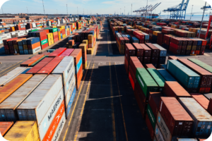 Vista aérea de um grande pátio de contêineres coloridos em um terminal portuário, representando a complexidade da logística e o vasto estoque disponível em um centro de distribuição. A imagem enfatiza a escala do transporte marítimo e a necessidade de planejamento logístico robusto para o interior do Amazonas.