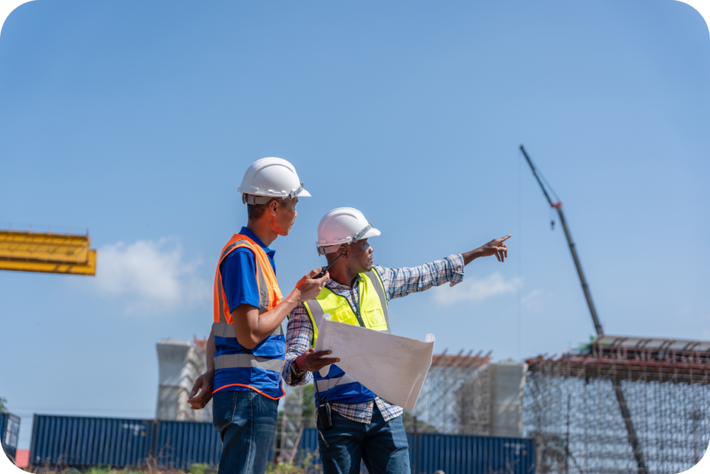 A imagem mostra dois homens, provavelmente engenheiros ou mestres de obra, em um canteiro de obras em um dia ensolarado. O homem à esquerda está de costas para o sol e veste uma camisa polo azul e laranja, colete de segurança laranja refletivo e capacete de segurança branco. O homem à direita, que parece estar falando no rádio ou celular, veste uma camisa xadrez, colete de segurança amarelo neon refletivo e capacete branco. Ele aponta para o horizonte, enquanto segura uma planta ou papel dobrado na outra mão. Ao fundo, sob um céu azul claro, vê-se a estrutura incompleta de uma construção, guindastes e contêineres de transporte, indicando que a imagem foi tirada em um local de construção industrial ou de infraestrutura de grande escala.
