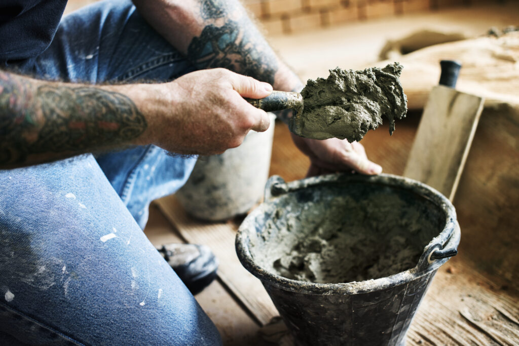 Homem com braços tatuados, usando calça jeans com respingos de tinta, agachado em uma obra enquanto utiliza uma colher de pedreiro para retirar massa de cimento de um balde preto. Ao fundo, detalhes de uma parede de tijolos em construção.