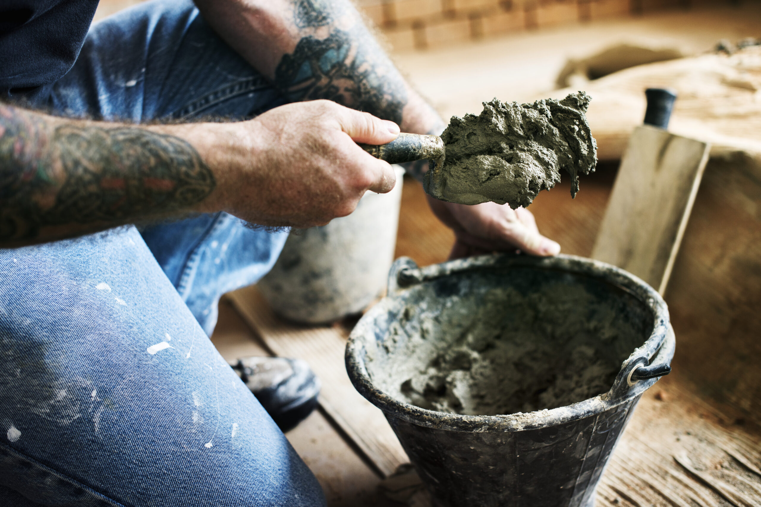 Homem com braços tatuados, usando calça jeans com respingos de tinta, agachado em uma obra enquanto utiliza uma colher de pedreiro para retirar massa de cimento de um balde preto. Ao fundo, detalhes de uma parede de tijolos em construção.