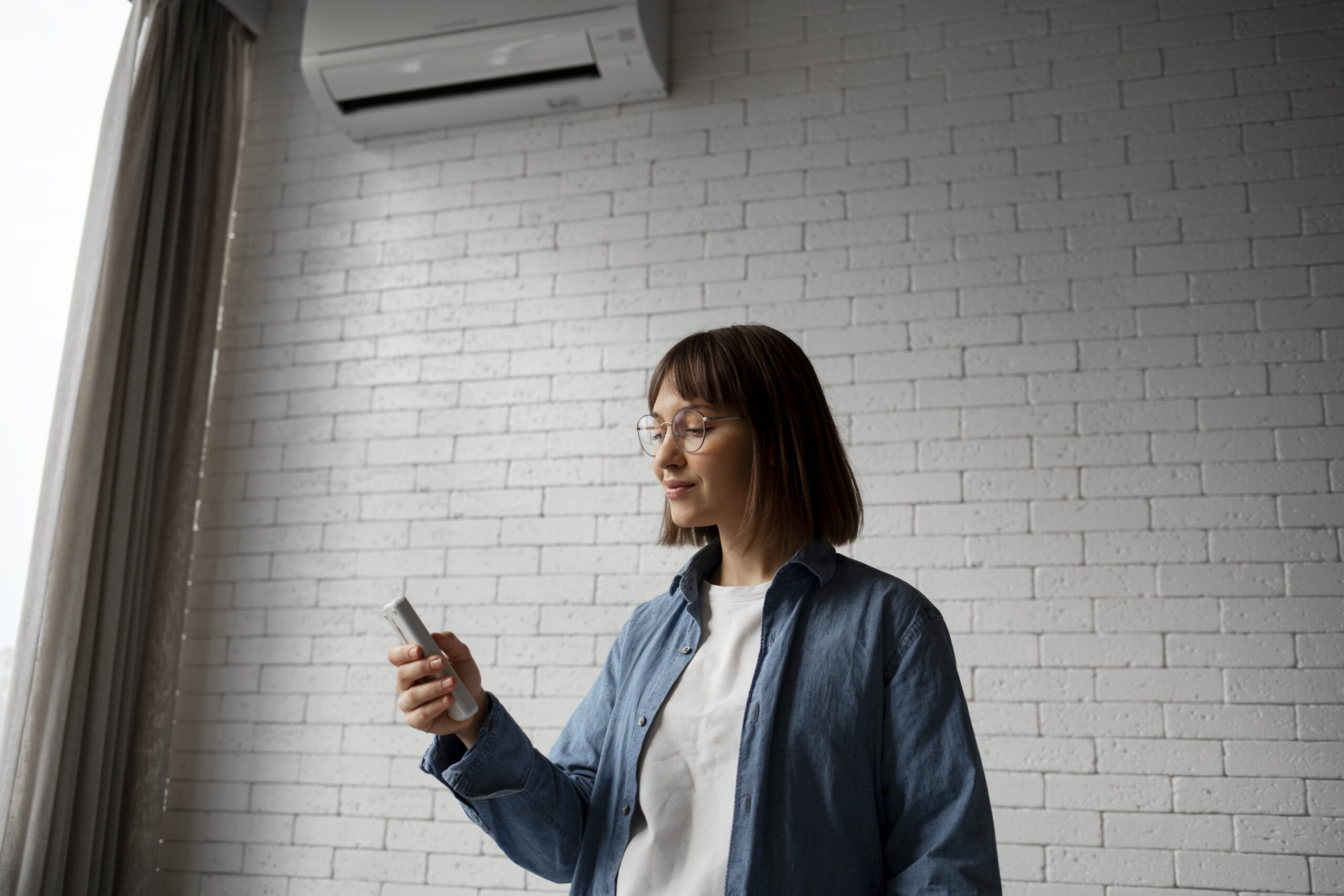 Mulher jovem com óculos usando um controle remoto para ajustar um aparelho de ar-condicionado instalado em uma parede de tijolos brancos, em um ambiente interno iluminado.