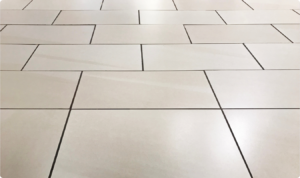 A imagem mostra um piso de cerâmica de cor creme em perspectiva de ângulo baixo, destacando o contraste entre as peças e o rejunte escuro. As peças retangulares estão dispostas em um padrão desencontrado (amarração), refletindo uma luz suave que sugere um acabamento acetinado ou semi-brilhante.