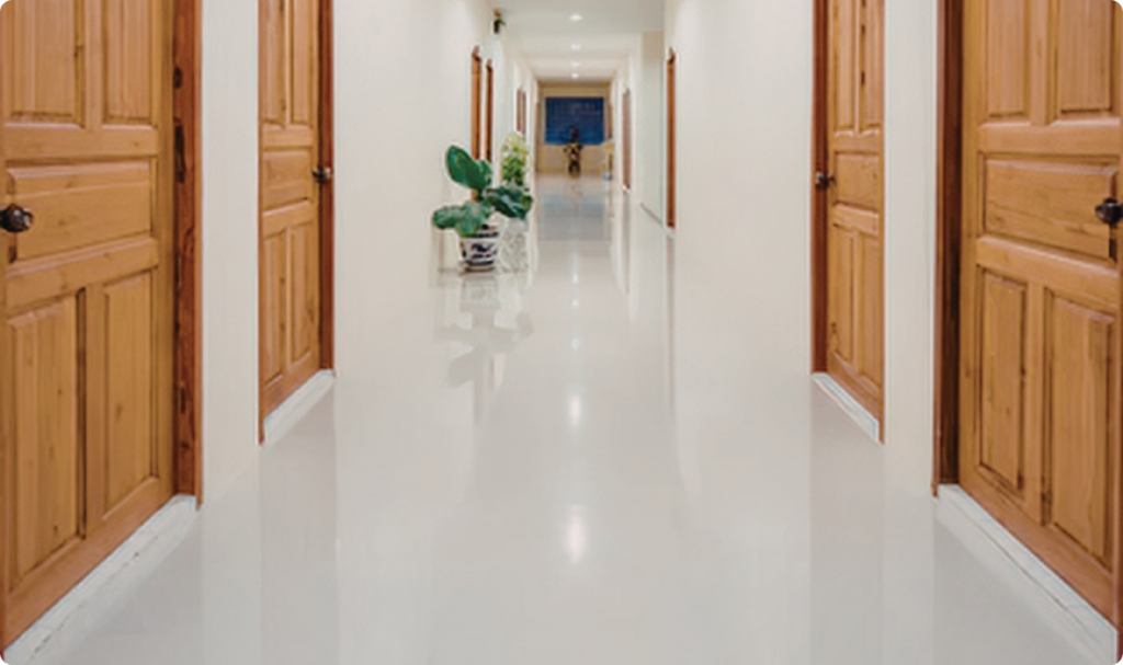 A imagem apresenta um corredor longo e iluminado com um piso cerâmico branco de alto brilho, que reflete intensamente as paredes e as portas de madeira. O ambiente possui paredes claras e várias portas de madeira em tons quentes distribuídas ao longo das laterais, criando um contraste entre a luminosidade do chão e a rusticidade da madeira.