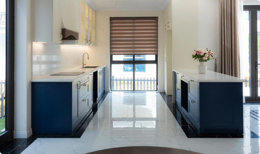 Cozinha moderna com armários em azul-marinho e bancadas brancas. O destaque é o piso de porcelanato branco polido com veios acinzentados, que possui um alto brilho e reflete a luz natural vinda de uma grande janela ao fundo. O design combina o estilo clássico das portas almofadadas com a sofisticação do chão espelhado e detalhes em preto nas bordas do piso.