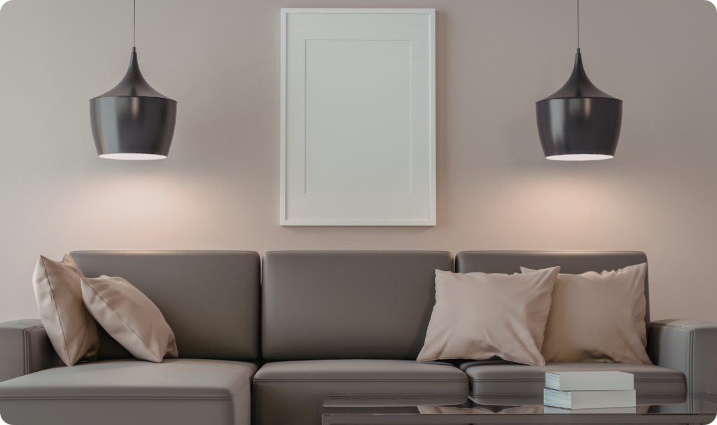Sala de estar minimalista com parede em tom bege neutro, decorada com um quadro de moldura branca centralizado e dois pendentes modernos de metal preto com iluminação focal quente. O ambiente inclui um sofá cinza de linhas retas com almofadas em tons de areia, criando uma composição elegante baseada em cores sóbrias e iluminação aconchegante.