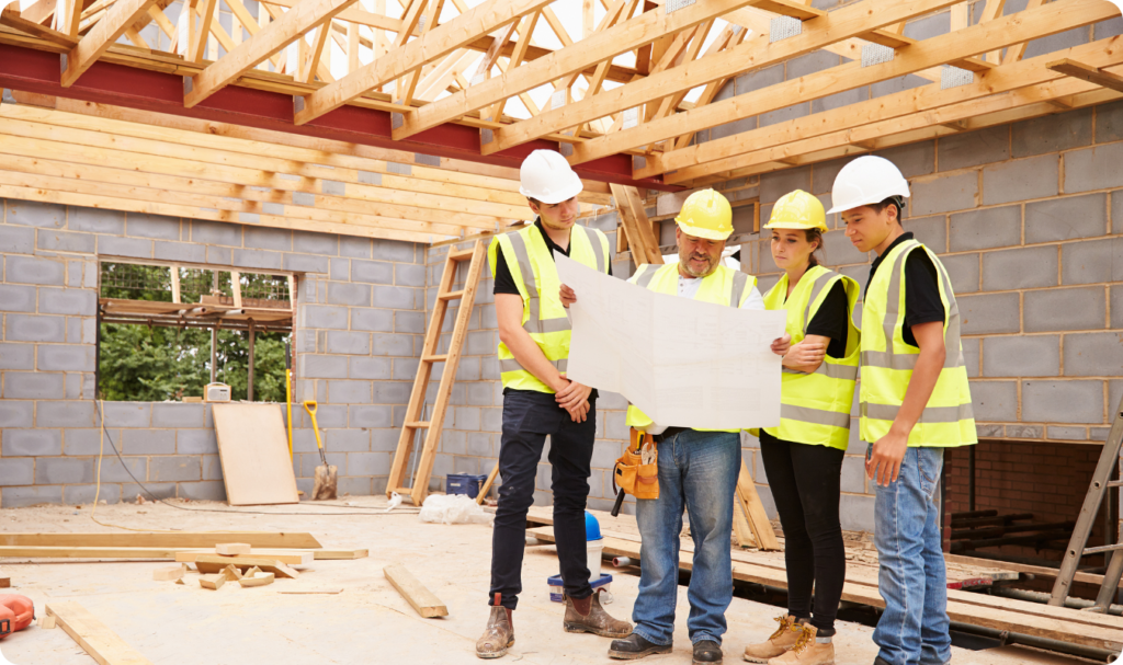 Equipe de construção civil, composta por quatro profissionais usando capacetes e coletes de segurança, analisando uma planta de engenharia em um canteiro de obras. Ao fundo, observa-se a estrutura interna de uma casa em fase de alvenaria com blocos de concreto e o madeiramento do telhado aparente, simbolizando a fase de planejamento técnico e infraestrutura.