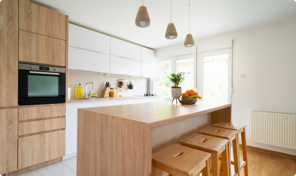 Cozinha moderna e iluminada com conceito aberto, apresentando uma ilha central de madeira clara com banquetas combinando. O ambiente destaca armários planejados em tons de branco e madeira, bancada de quartzo clara e iluminação pendente decorativa, transmitindo sofisticação e funcionalidade.
